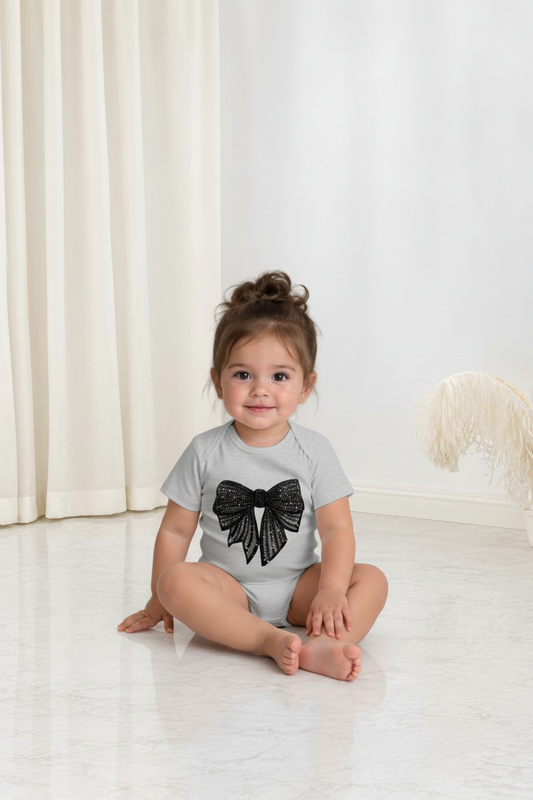 Bébé portant une grenouillère grise Le Cocon d'Or en coton avec un grand nœud noir à paillettes sur le devant
