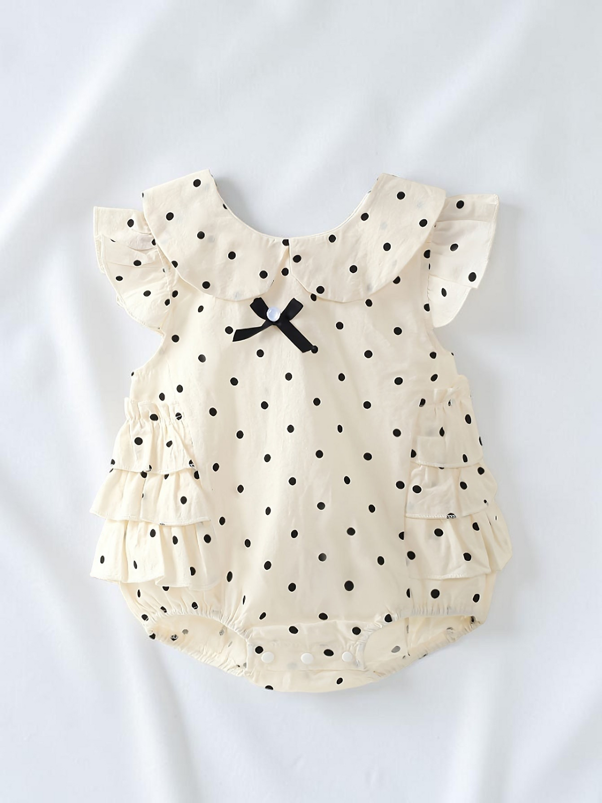Barboteuse Le Cocon d'Or en coton blanc à pois noirs, col Peter Pan, nœud décoratif et manches papillon pour bébé.