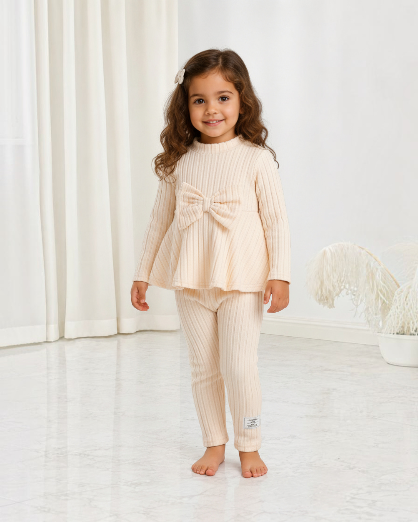 Petite fille souriante portant un ensemble Le Cocon d'Or en maille côtelée beige avec un haut à nœud papillon