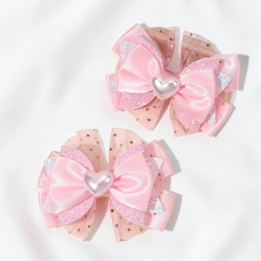Deux pinces à cheveux Le Cocon d'Or roses en forme de nœud avec un cœur central et des détails en tulle pailleté.