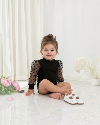 Bébé fille portant un body noir Le Cocon d'Or avec manches en dentelle à pois, assise sur un fond minimaliste.