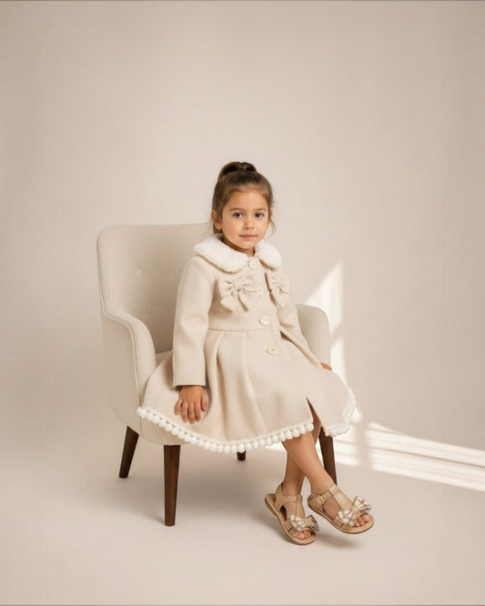 Petite fille portant le manteau Victoria Le Cocon d'Or beige avec col en dentelle, nœuds décoratifs et pompons.